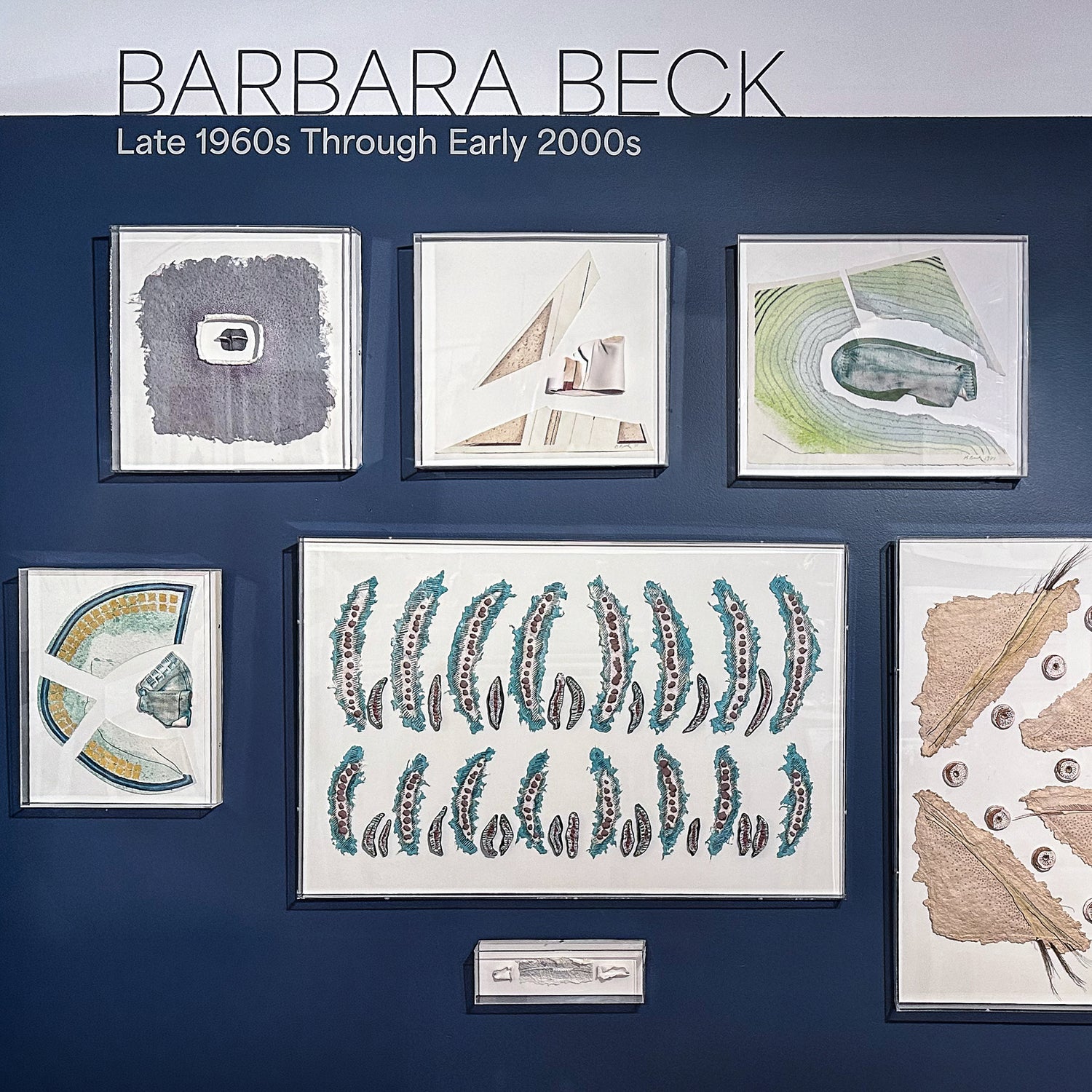 Collection of framed artworks by Barbara Beck from the late 1960s to early 2000s on a blue wall.