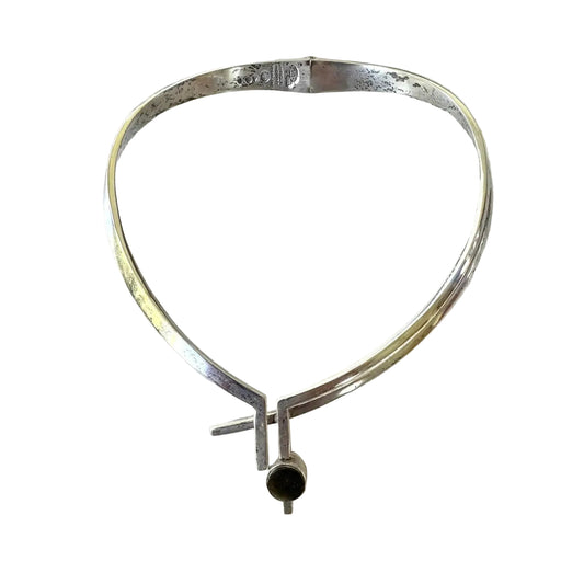 A polished silver-toned metal cuff bracelet with a unique Y-shaped clasp and a small dark circular accent at the bottom, displayed on a light textured surface.