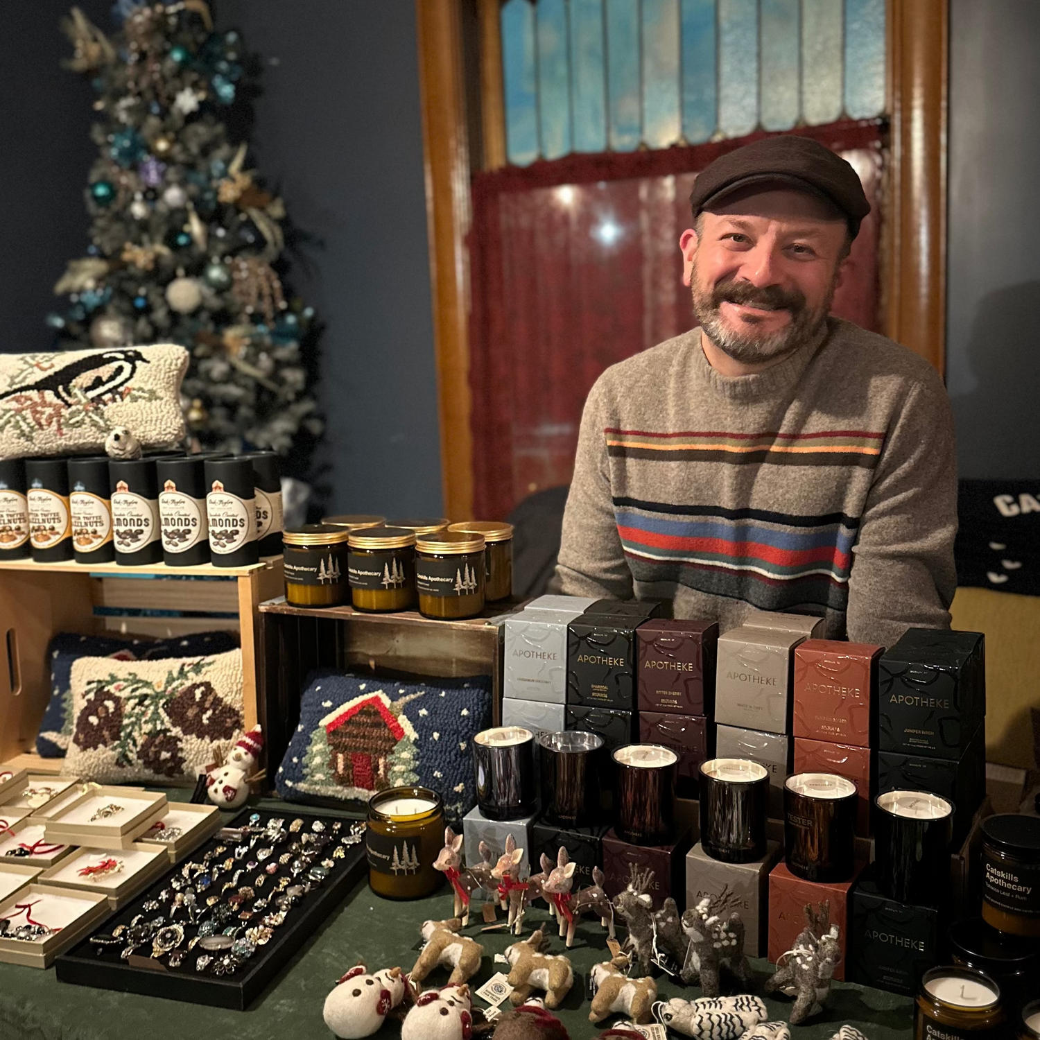 Man standing behind a table filled with various items including bottles and small objects, with a decorated Christmas tree in the background.